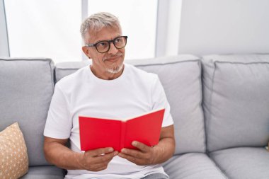 Orta yaşlı, kır saçlı, kitap okuyan adam evdeki kanepede oturuyor.