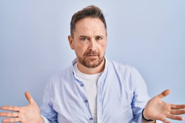 Middle age caucasian man standing over blue background clueless and confused with open arms, no idea concept. 
