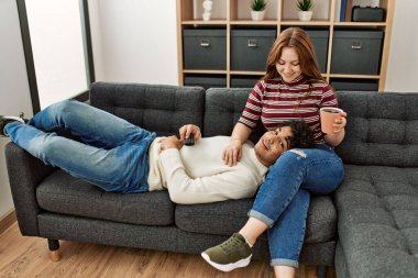 Mutlu gülümseyen genç çift evdeki kanepede oturup kahve içiyorlar..