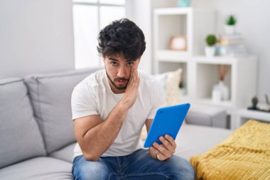 Hispanic man with beard using touchpad sitting on the sofa hand on mouth telling secret rumor, whispering malicious talk conversation 