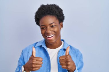 African american woman standing over blue background success sign doing positive gesture with hand, thumbs up smiling and happy. cheerful expression and winner gesture. 