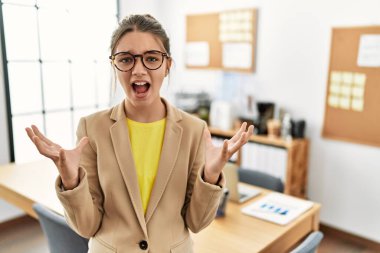 Young brunette teenager wearing business style at office crazy and mad shouting and yelling with aggressive expression and arms raised. frustration concept. 