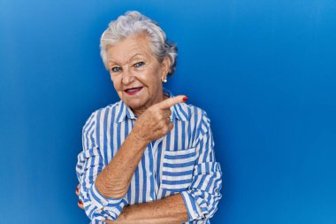 Mavi arka planda gri saçlı yaşlı bir kadın yüzünde kocaman bir gülümseme, eli ve parmağı kameraya bakıyor.. 