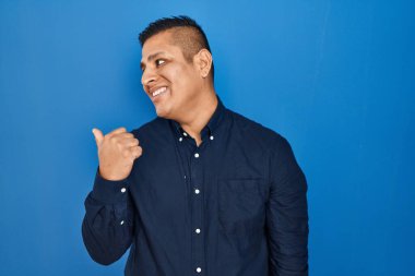 Hispanic young man standing over blue background smiling with happy face looking and pointing to the side with thumb up. 