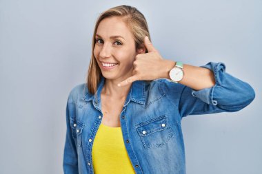 Mavi arka planda durup gülümseyen, el ve parmaklarıyla telefonla konuşan genç sarışın kadın. iletişim kavramları. 