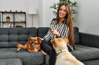 Genç İspanyol kadın akıllı telefon kullanıyor. Kanepede köpeklerle oturuyor.