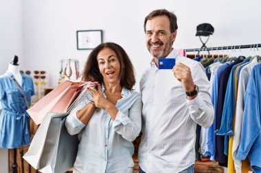 Hispanic middle age couple holding shopping bags and credit card with a big smile on face, pointing with hand finger to the side looking at the camera. 