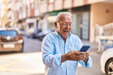 Yaşlı, gri saçlı, kendine güvenen, caddede akıllı telefon kullanan bir adam.
