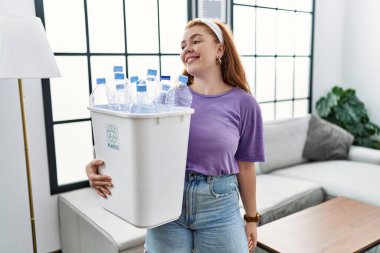 Geri dönüşüm sepetini elinde plastik şişelerle elinde tutan genç kızıl saçlı kadın yüzünde gülümseme ve doğal bir ifadeyle yan yana bakıyor. Kendinden emin gülen. 