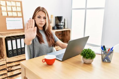 Young brunette woman working at the office with laptop doing stop sing with palm of the hand. warning expression with negative and serious gesture on the face. 