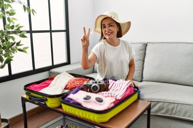 Beautiful middle age hispanic woman packing summer clothes in suitcase smiling looking to the camera showing fingers doing victory sign. number two. 