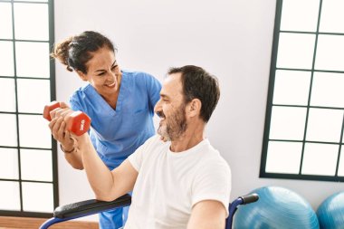 Orta yaşlı bir adam ve kadın, fizyoterapi kliniğinde tekerlekli sandalyede oturan dambılı kullanarak rehabilitasyon seansı yapıyorlar.