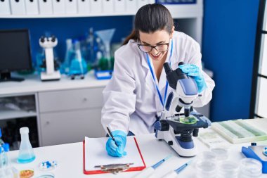 Young hispanic woman scientist write on document using microscope at laboratory