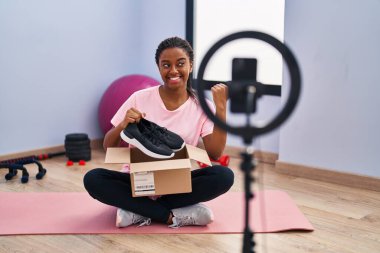 Örgülü genç Afro-Amerikalı spor salonunda akıllı telefonlu vlog dersleri kaydediyor. Başparmağını yan tarafa uzatıyor ve ağzı açık gülümsüyor. 