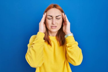 Mavi arka planda duran genç bir kadın, elleri başının üstünde, stresten baş ağrısı çekiyor. migren ağrısı. 