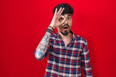 Young hispanic man with beard standing over red background doing ok gesture shocked with surprised face, eye looking through fingers. unbelieving expression. 