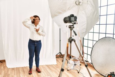 Orta yaşlı İspanyol kadın fotoğraf stüdyosunda modellik yapıyor gülümsüyor ve mutlu yüzlü eller ve parmaklarla çerçeve yapıyor. yaratıcılık ve fotoğrafçılık kavramı. 