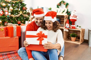 Orta yaşlı İspanyol çift Noel şapkası takıyor. Evde yerde oturup hediyeyi açmak..