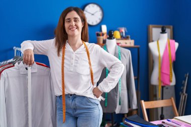 Young woman tailor smiling confident leaning on clothes rack at sewing studio