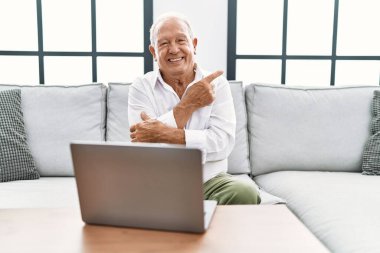 Senior man using laptop at home sitting on the sofa with a big smile on face, pointing with hand and finger to the side looking at the camera. 