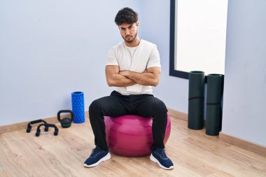 Hispanic man with beard sitting on pilate balls at yoga room skeptic and nervous, disapproving expression on face with crossed arms. negative person. 