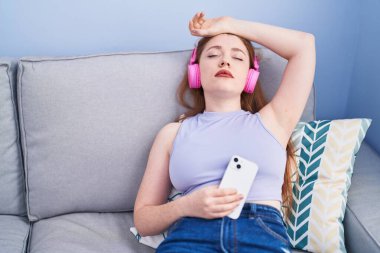 Genç kızıl saçlı kadın evdeki kanepede rahat bir şekilde müzik dinliyor.
