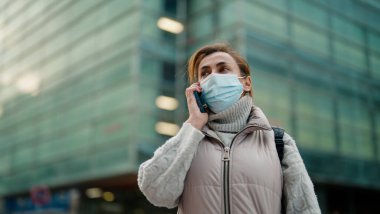 Orta yaşlı, tıbbi maske takan bir kadın sokakta akıllı telefondan konuşuyor.