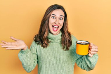 Young hispanic girl drinking a cup of coffee celebrating achievement with happy smile and winner expression with raised hand 