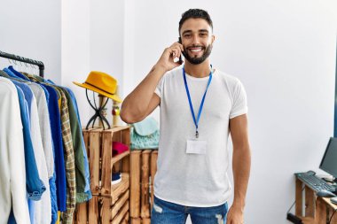 Genç Arap esnaf akıllı telefondan konuşuyor giyim mağazasında elbise rafının yanında duruyor.