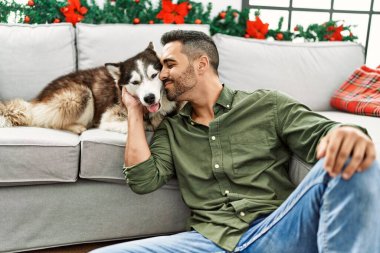 Young hispanic man kissing dog sitting on sofa by christmas decor at home
