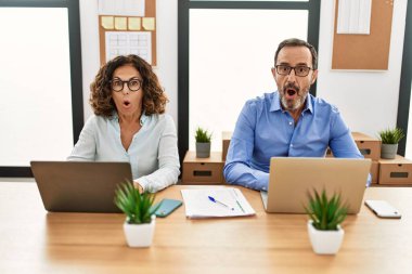 Middle age hispanic woman and man sitting with laptop at the office afraid and shocked with surprise expression, fear and excited face. 