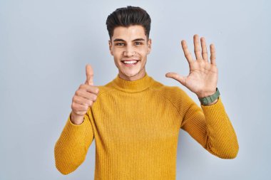 Young hispanic man standing over blue background showing and pointing up with fingers number six while smiling confident and happy. 