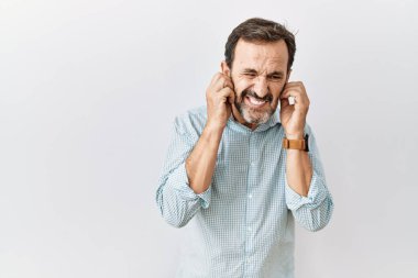 Middle age hispanic man with beard standing over isolated background covering ears with fingers with annoyed expression for the noise of loud music. deaf concept. 