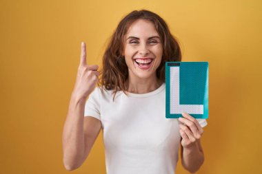 Beautiful brunette woman holding l sign for new driver surprised with an idea or question pointing finger with happy face, number one 