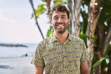 Genç İspanyol adam deniz kenarında yürürken kendinden emin gülümsüyor.