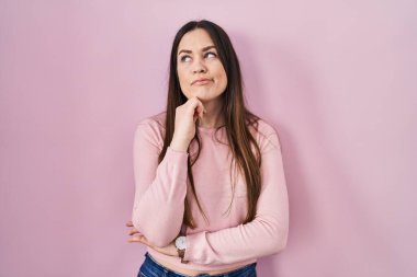 Pembe arka planda eli çenesinde duran genç esmer kadın soru sormayı, düşünceli yüz ifadesini düşünüyor. Düşünceli bir yüzle gülümsüyor. şüphe kavramı. 