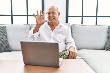 Senior man using laptop at home sitting on the sofa showing and pointing up with fingers number five while smiling confident and happy. 