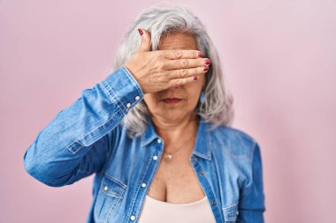 Middle age woman with grey hair standing over pink background covering eyes with hand, looking serious and sad. sightless, hiding and rejection concept 