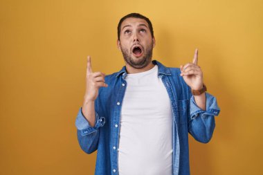 Hispanic man standing over yellow background amazed and surprised looking up and pointing with fingers and raised arms. 