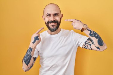 Young hispanic man with tattoos standing over yellow background smiling cheerful showing and pointing with fingers teeth and mouth. dental health concept. 