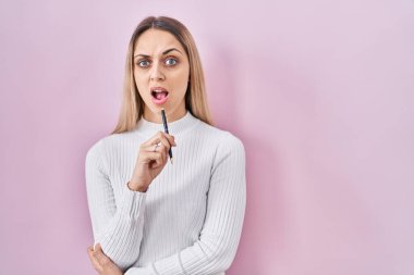 Pembe arka planda elinde kalem tutan genç sarışın kadın. Hiçbir şey bilmiyor ve kafası karışık. şüphe kavramı. 