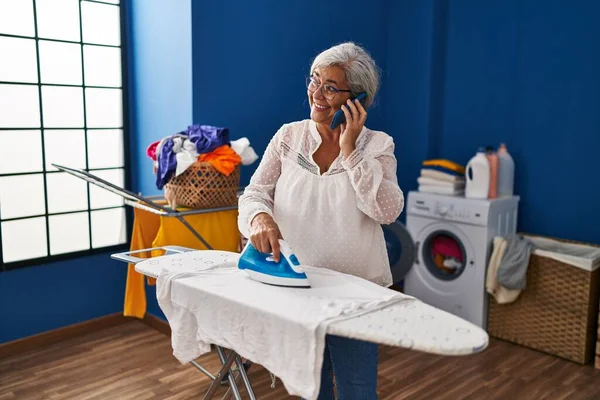 洗濯室でスマートフォンのアイロン掛け服に話している中年女性 — ストック写真