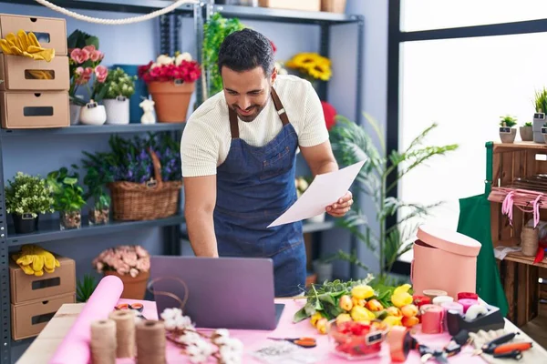 Young hispanic man florist using laptop reading paper at florist