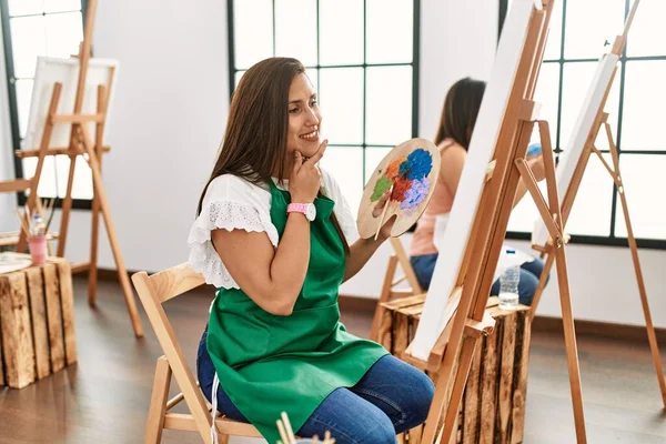 若いヒスパニック系のアーティストの女性は 集中的な表現について考えて顎を手でアートスタジオでキャンバスに絵を描く 笑顔で思いやりのある顔 疑わしい概念 — ストック写真