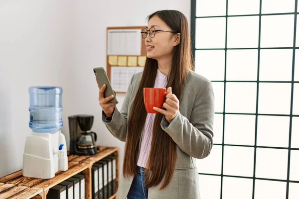 Young chinese businesswoman using smartphone and drinking coffee at the office.