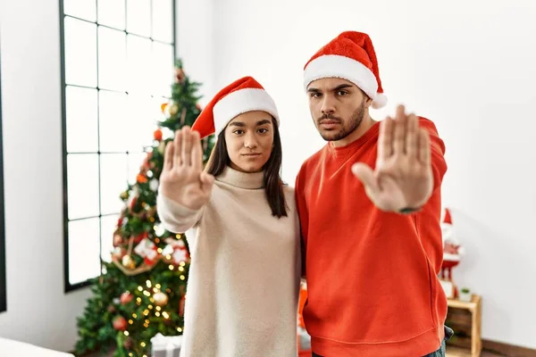 クリスマスツリーのそばに立つ若いヒスパニック系のカップルは 手の手のひらで歌うのを停止します 顔に負の重大なジェスチャーで — ストック写真
