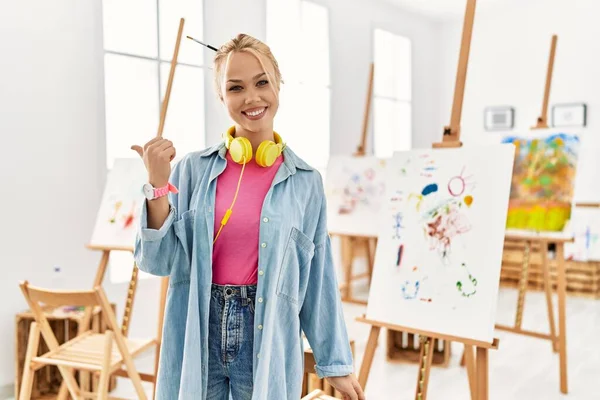 Young caucasian girl at art studio smiling with happy face looking and pointing to the side with thumb up.