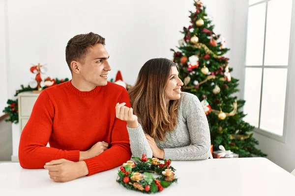Young hispanic couple sitting at the table on christmas looking away to side with smile on face, natural expression. laughing confident.