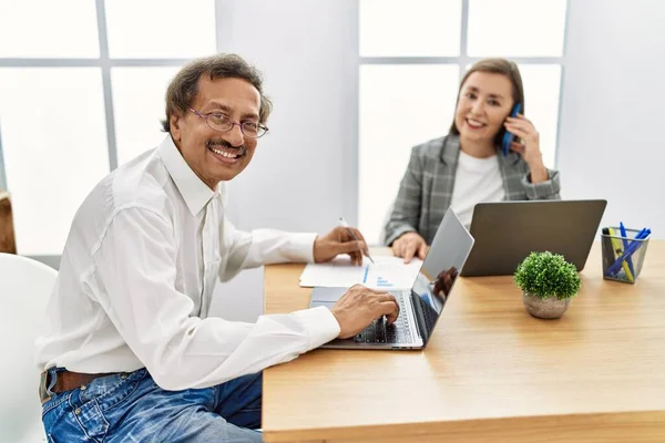 Middle age man and woman business workers using laptop and talking on the smartphone at office