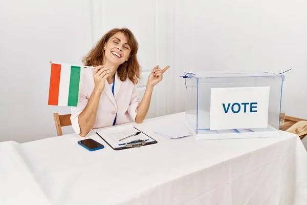 Beautiful caucasian woman at political campaign election holding hungary flag smiling happy pointing with hand and finger to the side